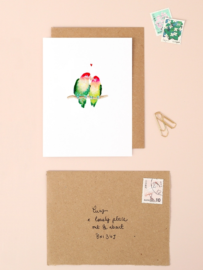 An image of a mini, rectangular white greetings card with an illustration of a couple of parrots kissing with tiny red heart above their heads. The card is seen on top of a brown rectangular kraft envelope.