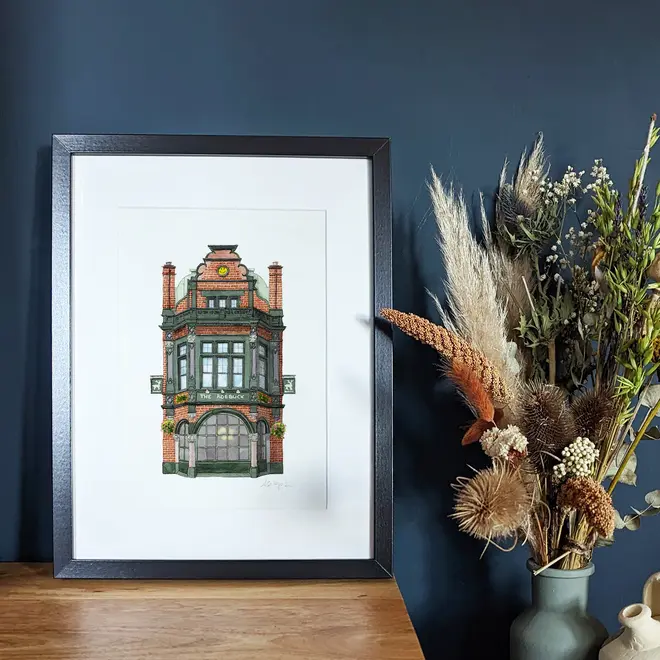 Borough The Roebuck Pub Giclée Print (Unframed), a print of a pub sitting in a black frame on a wooden surface against a navy blue backdrop. It is accompanied by some flowers. 