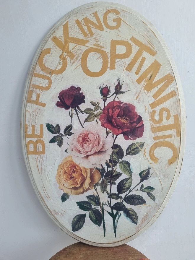 An art panel made from an upcycled table top. The image is vintage roses on a cream background with text that reads 'be fucking optimistic' in mustard yellow..