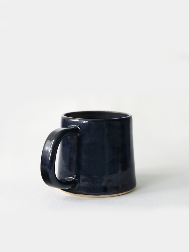 A handmade ceramic standard mug in glossy jet black glaze with a chunky loop handle, photographed against a plain light grey background