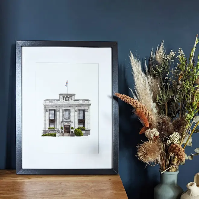 Islington Town Hall Giclée Print (Unframed), a print of a town hall sitting in a black frame on a wooden surface against a navy blue backdrop. 