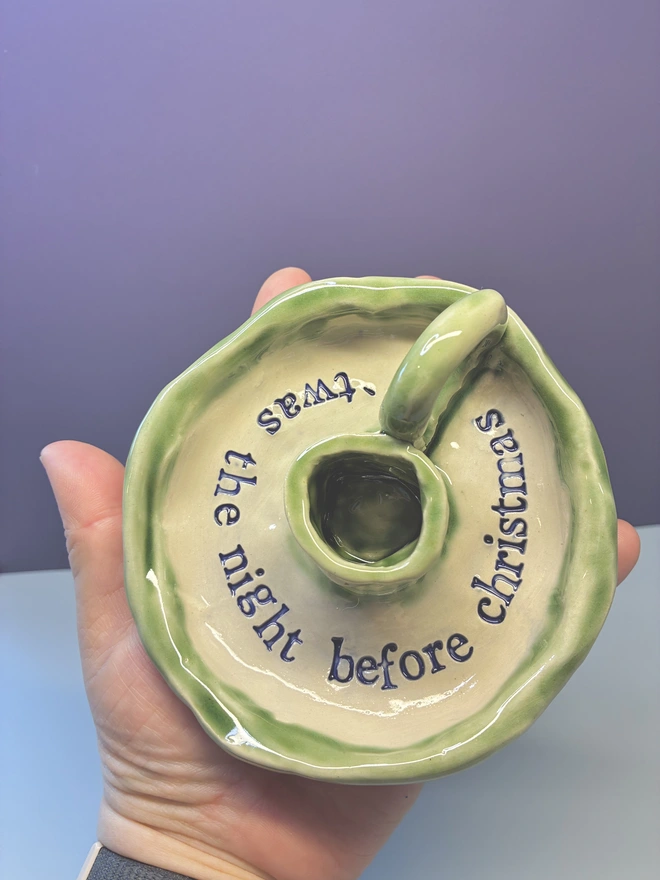 Handmade ceramic Christmas taper candlestick holder with a soft green glazed edge and the words “‘Twas the night before Christmas” in dark blue lettering, arranged around the candle holder opening. Photographed against a pale background.