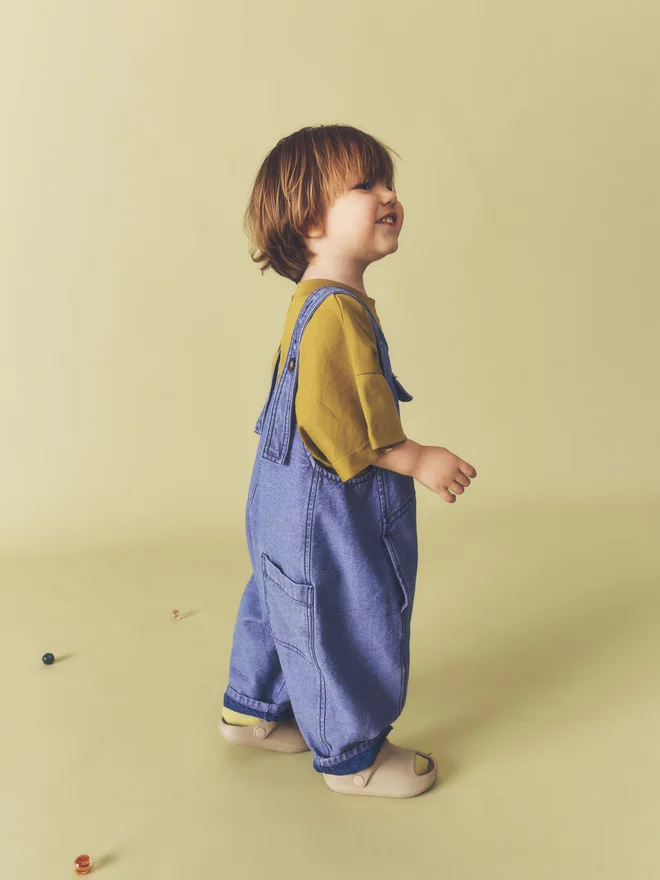 Side view of a toddler smiling against a pale yellow backdrop, wearing Studio Koter washed blue denim Kai dungarees with side patch pocket, over an ochre t-shirt.