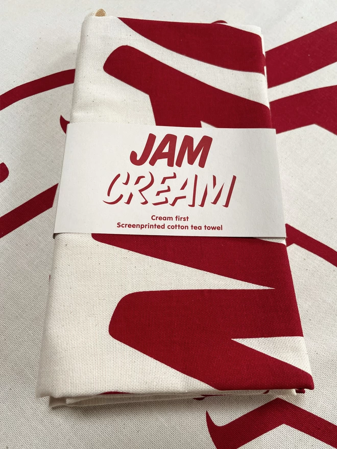 A Cream First tea towel folded up with a belly band around, and sat on another teatowel. It says Cream first, Screenprinted cotton teatowel in red ink on cream coloured card and cotton fabric.