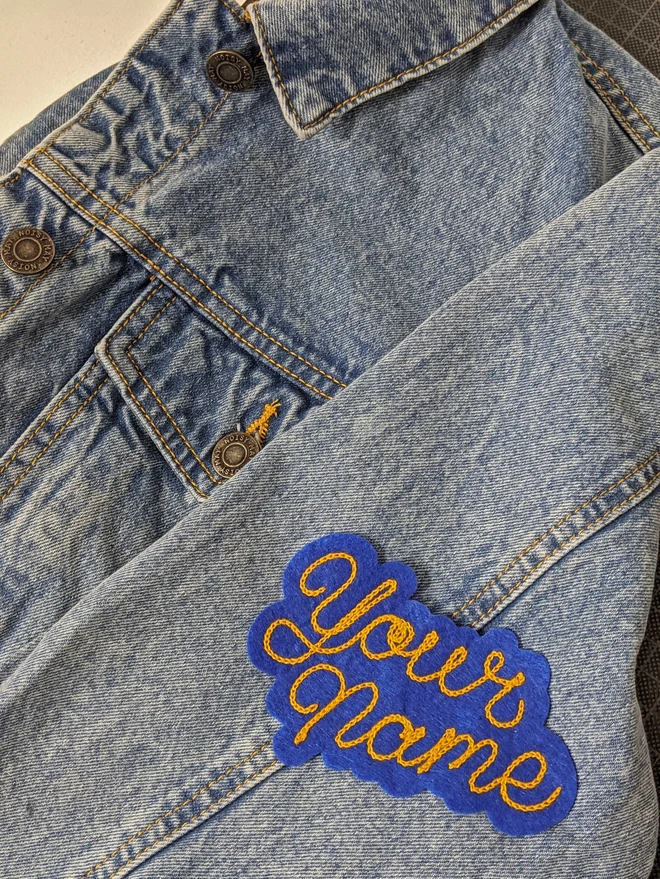 'Your Name' Patch, with Gold cursive embroidery on a navy blue backing felt, presented on a denim jacket. 