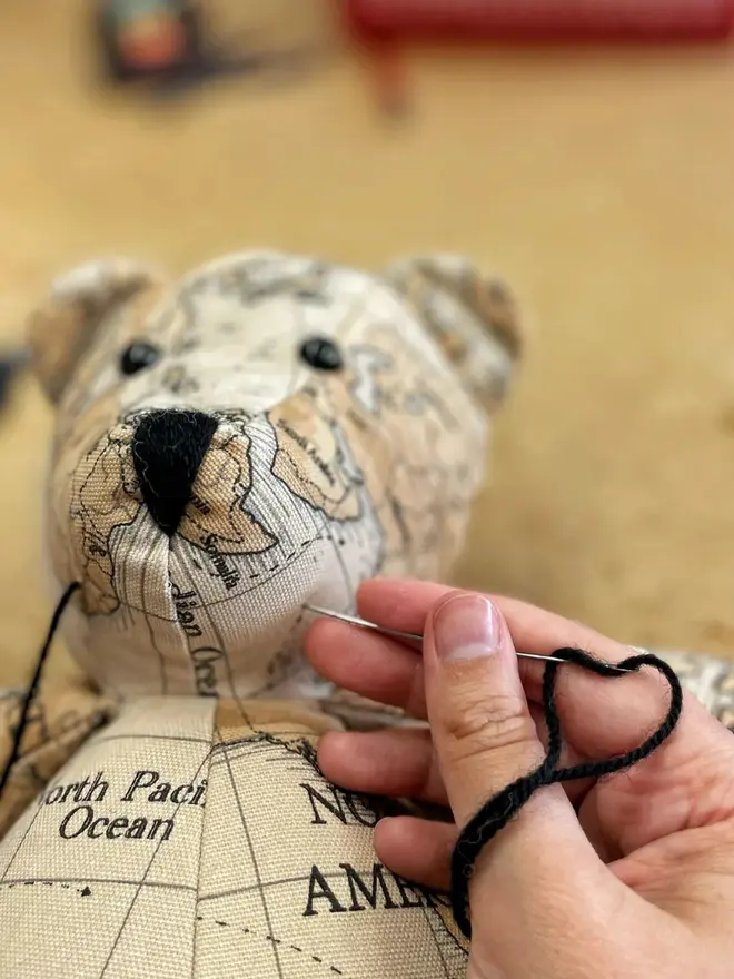 Teddy bear made from map-print fabric sitting upright on a wooden table, wearing a brown bow.