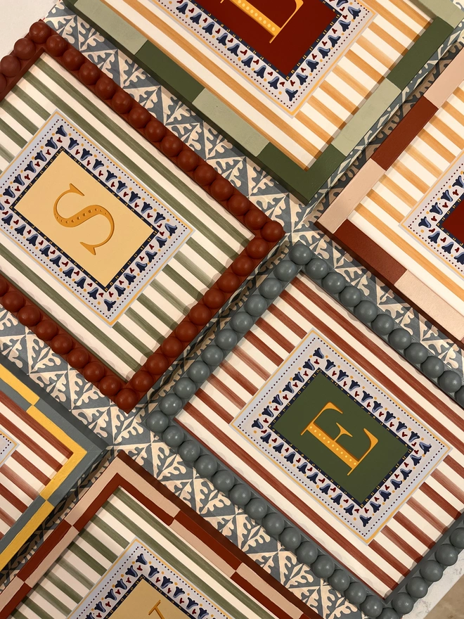 A collection of colourful hand painted bobbin and striped frames with letter prints inside surrounded by a striped border. Colourful wall art display by annabel wond interiors. 