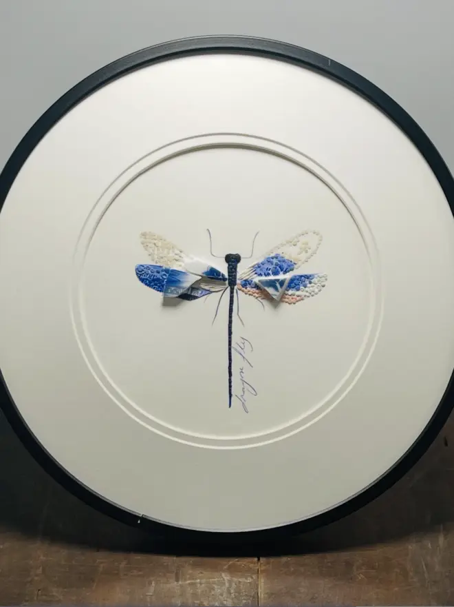 Hand-drawn, painted, mosaic clay, blue vintage Willow Tree pottery insect on white double mounted card, in a black circular frame.