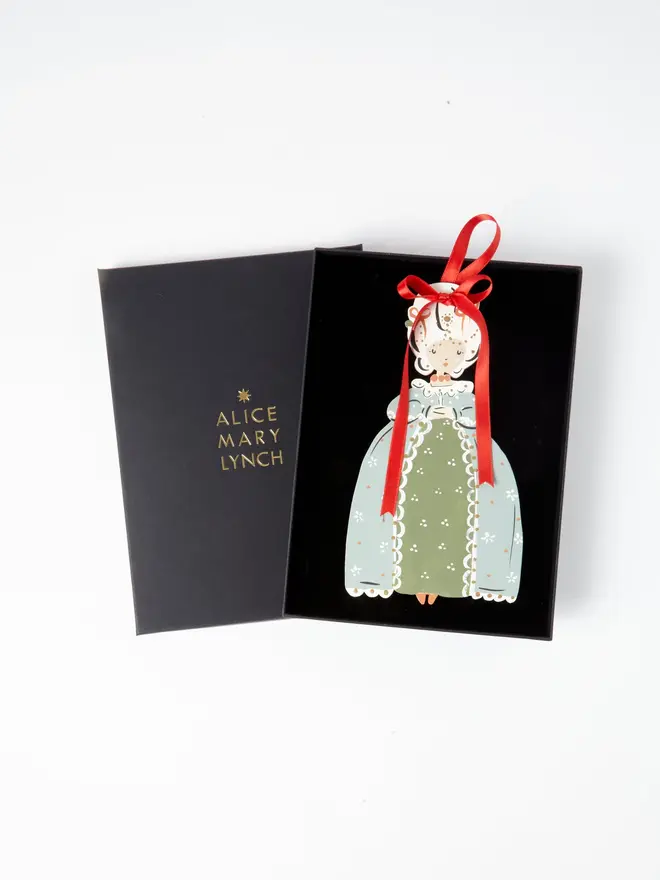 Sorrel Princess Tree Dancer. the sorrel princess tree dancer hanging ornament sits inside its black presentation box, with the lid open to reveal "alice mary lynch" embossed in gold lettering. the princess figure has an elaborate powdered wig decorated with pearls and jewels, wearing a sage green gown with an olive skirt panel and white lace trim, hanging by its red satin ribbon bow. the branded box gives the piece a polished, gift-ready presentation.