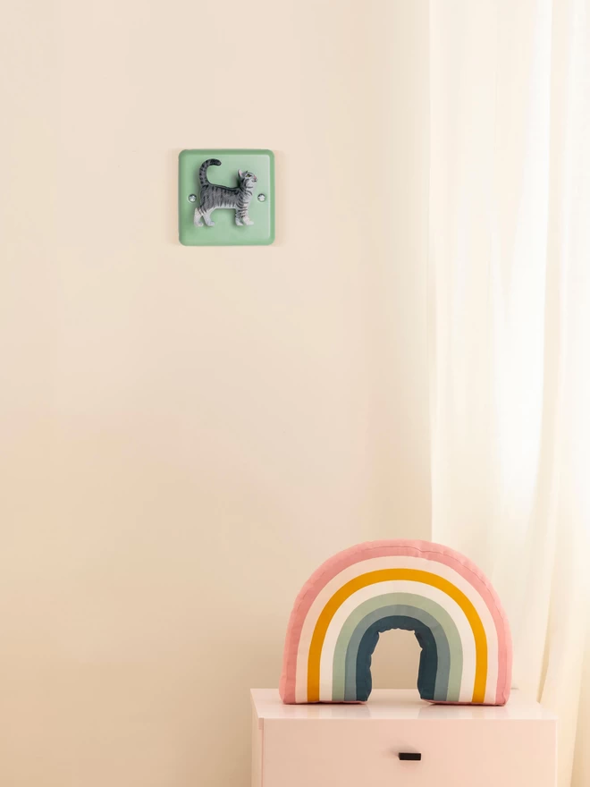 A pastel green dimmer light switch with a happy looking grey cat as the rotary knob to turn the lights on and off on a light pastel pink wall. The light switch plate is beryl green and made of metal, epoxy coated steel by varilight. The grey cat is made of plastic. The animal light switch brand is Candy Queen Designs. There is a small chest of drawers in the foreground with a pastel coloured rainbow sitting on it made from soft cotton material.