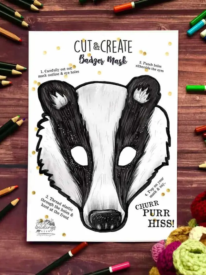 Cut And Create Forest Animals Children’s Masks Kit, an illustrated raccoon face, surrounded by coloured pencils on a dark wood background.
