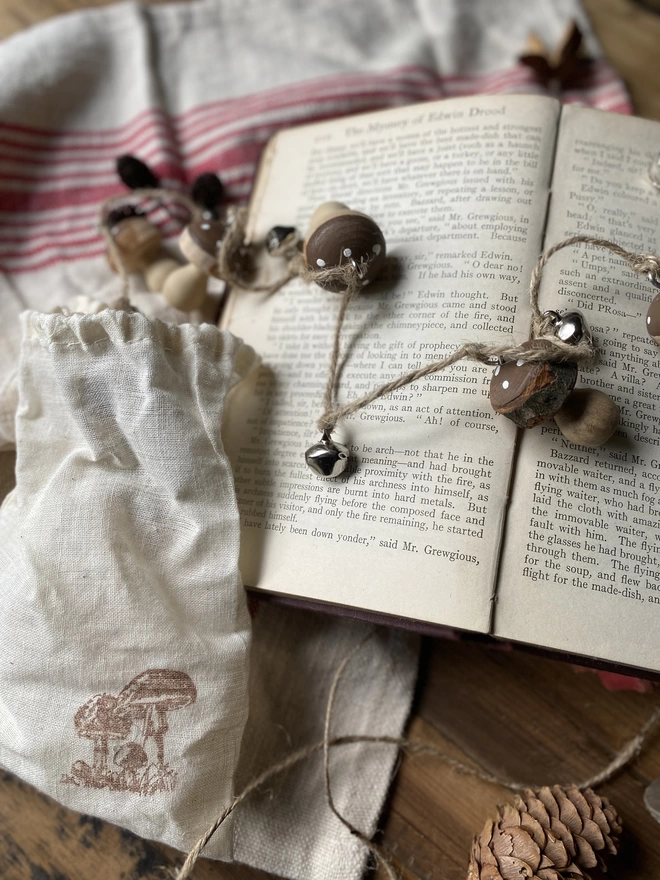 A string of Hand Painted Wooden Toadstool Bell Garlands trailing across an opened book atop a red and white striped cloth, on display with a small white pouch and some decorative pine cones