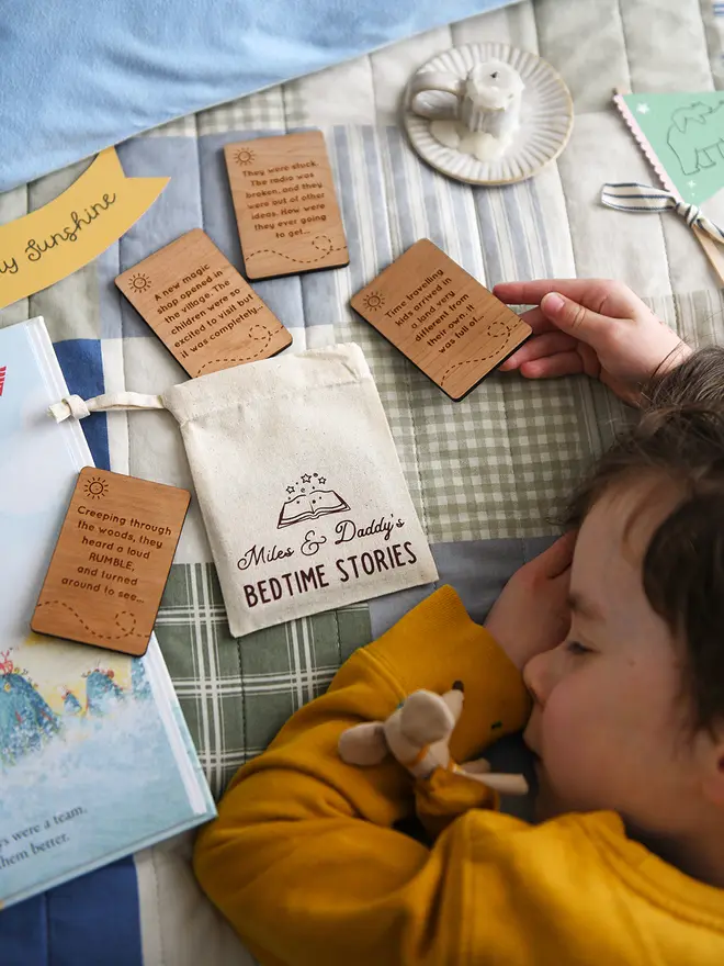 Personalised bedtime story set for dads and children featuring engraved wooden story prompt cards and a drawstring cotton bag reading 'Miles & Daddy’s Bedtime Stories', displayed on a patchwork quilt with a soft toy, picture book, and a young child sleeping beside it.