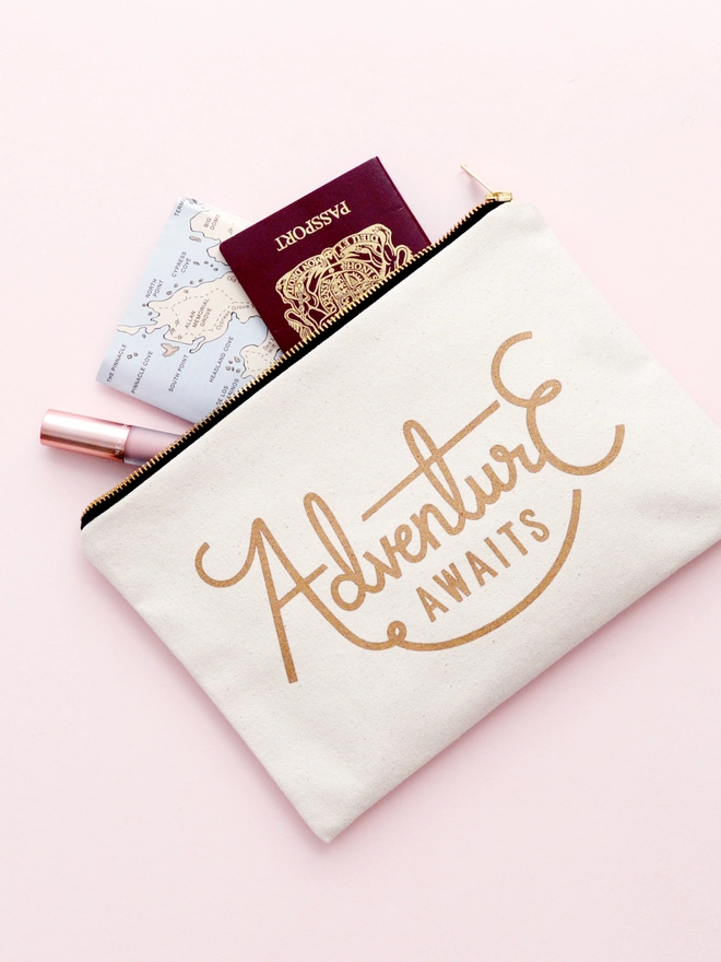 a natural canvas zip pouch with the words adventure awaits on a pink background with a passport and map spilling out 