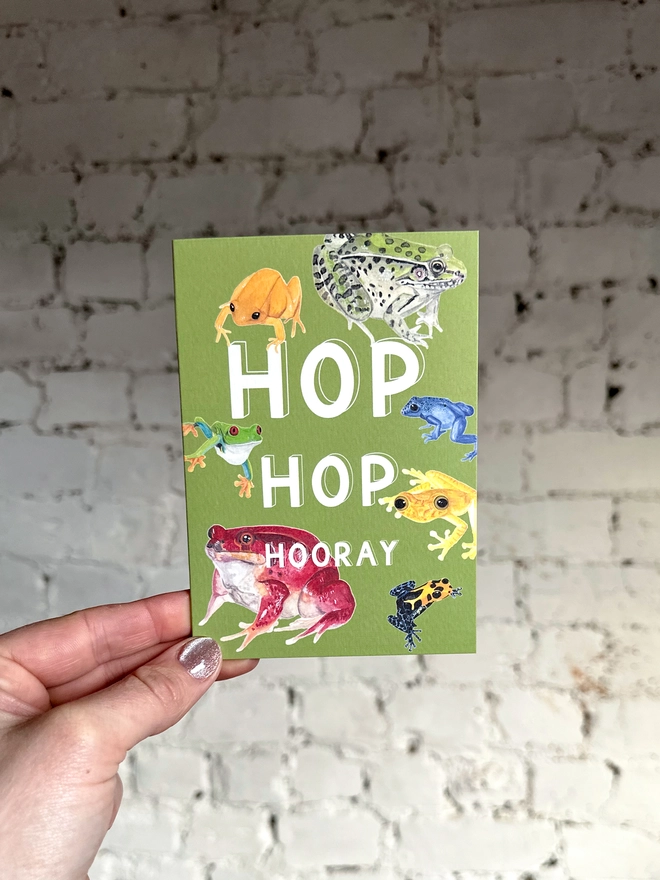 a green greetings card featuring a selection of frog illustrations with the text “hop hop hooray”