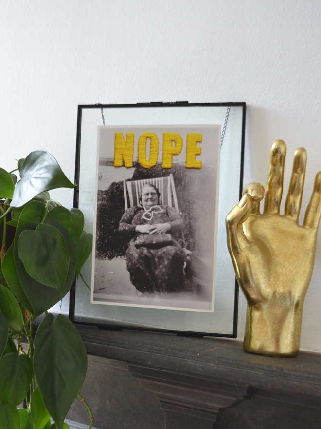  B & W photo print of woman with yellow embroidered ‘nope’ framed on mantlepiece 