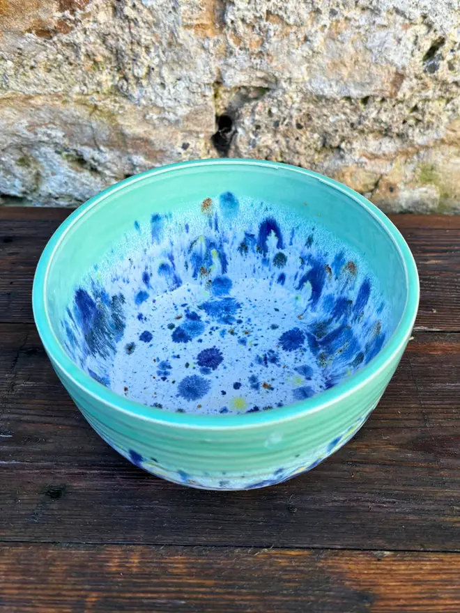 Shoreline Small Bowl, a colourful bowl placed on a wooden surface in front of a brick wall. 