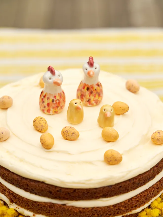 2 ceramic Easter chicks and 2 ceramic Easter chickens on top of a large carrot cake with mini eggs, on a cake stand surrounded by mimosa flowers on a stripy yellow and white tablecloth