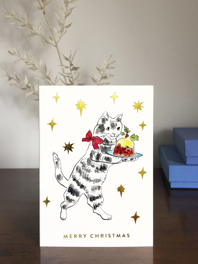 Christmas scene featuring a Christmas card with a cat carrying a Christmas Pudding surrounded by gold foil stars. 