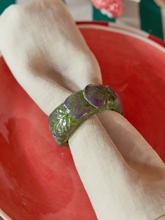 Detailed close-up of a handmade ceramic Brussels sprout napkin ring holding a folded linen napkin over a coral-red plate, showing the textured green and purple glazed leaves.