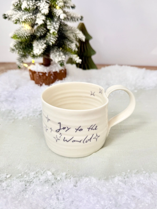 cream hand thrown porcelain mug with the words 'Joy to the World' printed onto the side with pretty line drawn stars. Sitting on a tabletop with snow, a mini lit Christmas tree 