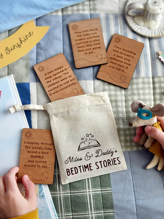 Personalised bedtime story set for dads and children featuring engraved wooden story prompt cards and a drawstring cotton bag reading 'Miles & Daddy’s Bedtime Stories', displayed on a patchwork quilt with a soft toy, picture book, and a young child holding one of the cards.