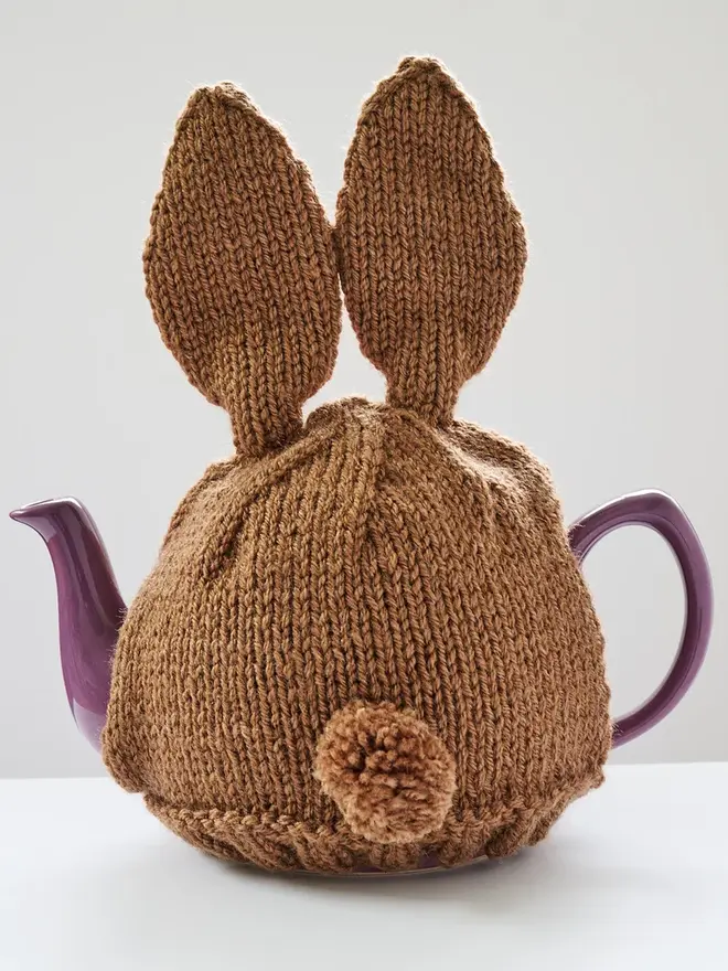 Hand Knitted Bunny Rabbit Tea Cosy, a rabbit bunny tea cosy sitting on a grey surface against a white backdrop. It fits over a purple tea pot. 