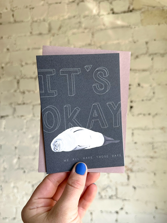 a dark grey greetings card featuring an illustration of a seal with the text “It’s Okay - we all have those days” with a muted purple envelope