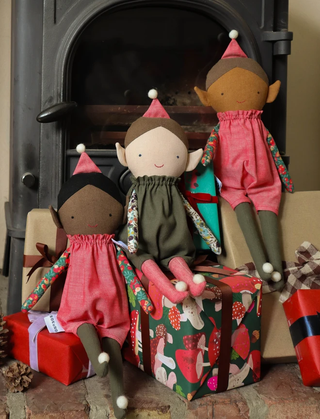 Three handmade Christmas elf dolls with pointed ears, wearing olive green romper suits with colourful floral Liberty print sleeves, red hats and matching long legs with white pom-pom feet. Light, mid and dark skin tones.