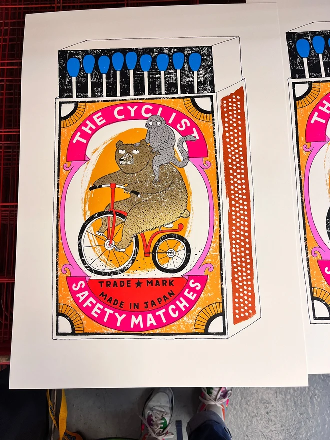 Bear On A Bike Matchbox Screenprint Big Cyclist's Burden, a colourful matchbox design in the centre of a white screenprint. It sits on a studio rack. 
