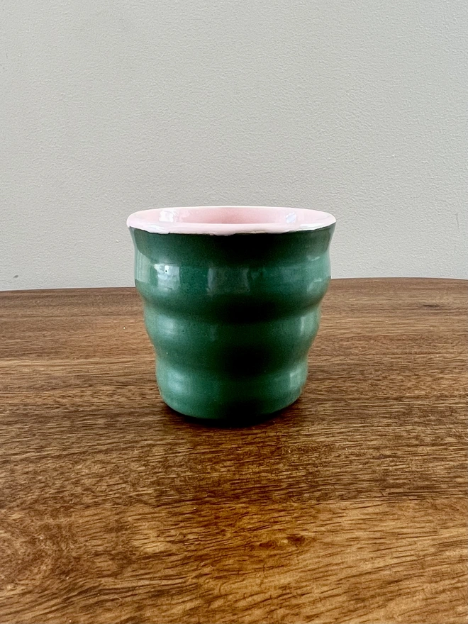 ceramic wavy tumbler with dark green glaze on the outside and light pink on the inside. the tumbler holds 250ml of liquid and sits on a wooden table