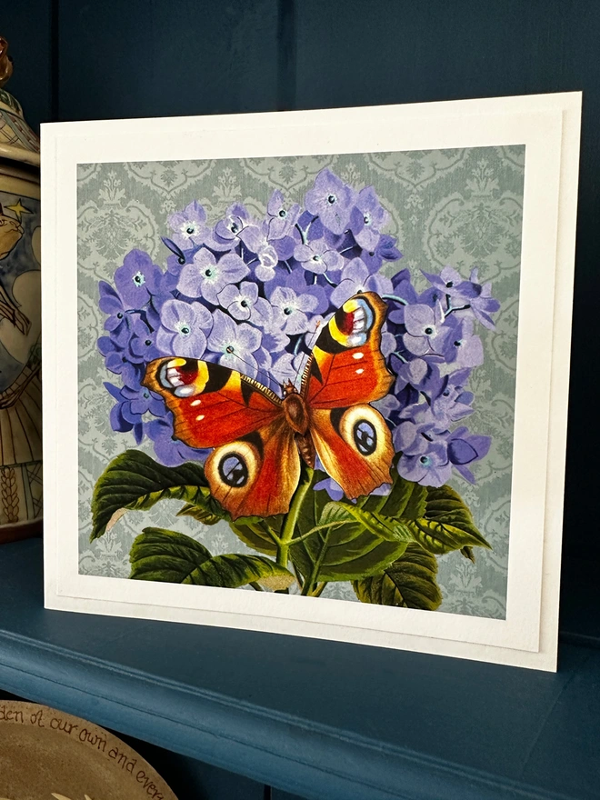 Blue hydrangea flower with a 3D paper cut Peacock butterfly on a teal backgrouund. Hand made card displayed oon a blue dresser in the home.