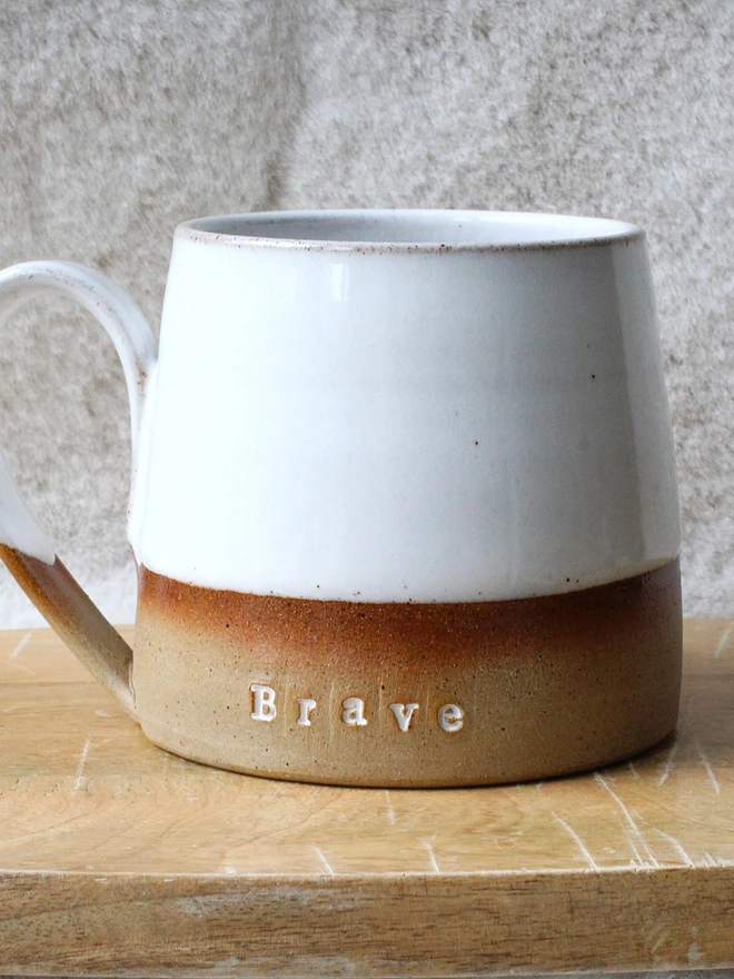Rustic ceramic mug with a white speckled upper glaze and brown lower half, featuring the word “Brave” on the front, sitting on a wooden table against a neutral background.