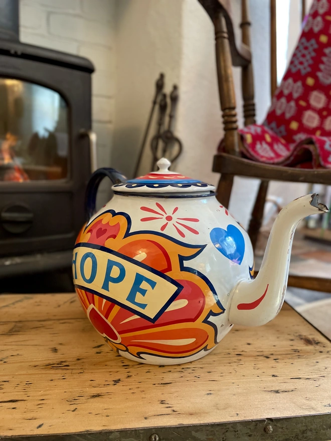 White vintage teapot, hand painted with a fairground inspired design in pink, orange, yellow and red. 'Hope' is painted in a banner across the centre.