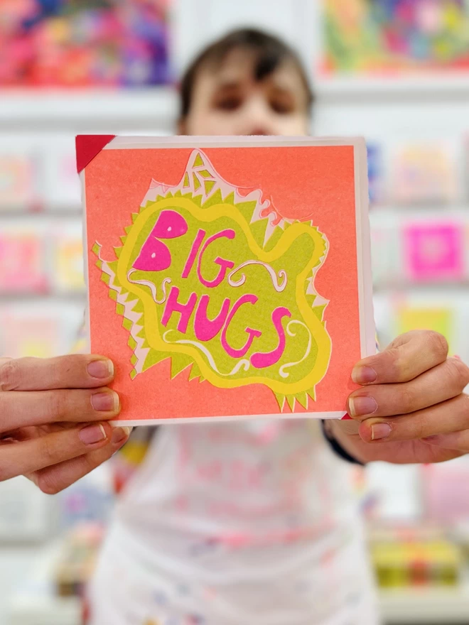 A bright & cArtist holding a colourful riso print get well soon card featuring the words Big Hugs in fluro pinkolourful riso print get well soon charity card featuring the words Big Hugs in fluro pink