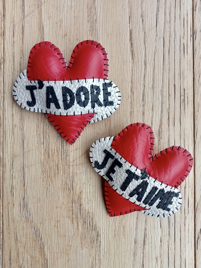 Two red heart brooches, laying on a wooden surface. The black lettering across the white scrolls say J'ADORE and JE T'AIME