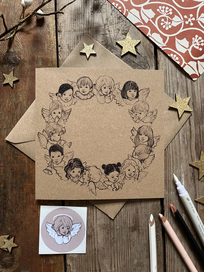 a kraft square Christmas gretting card featuring a circle wreath of angels printed in black drawn in a vintage story book style