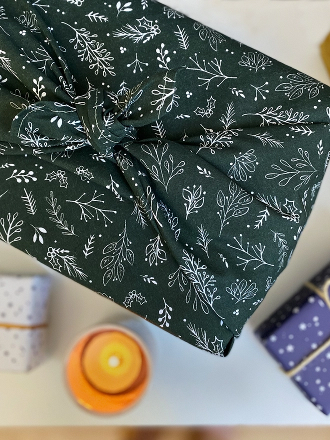 A gift wrapped in deep green cotton fabric wrap with a botanical design, is on a white desk beside other gifts and a candle.