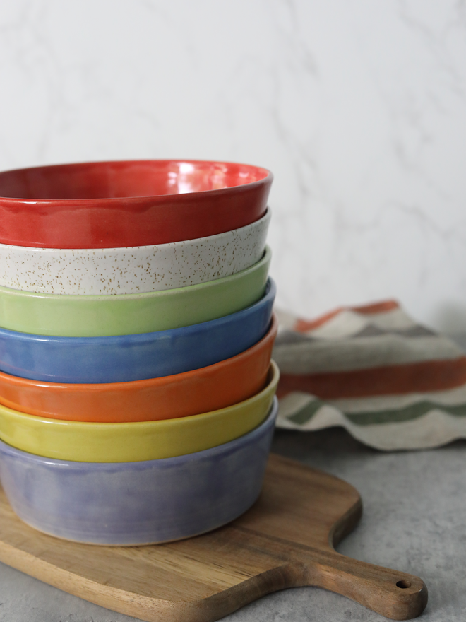 A tall stack of seven ceramic pasta bowls in rainbow colours — red, speckled white, green, blue, orange, yellow, and lavender — stacked on a wooden chopping board, with a striped linen cloth beside them.