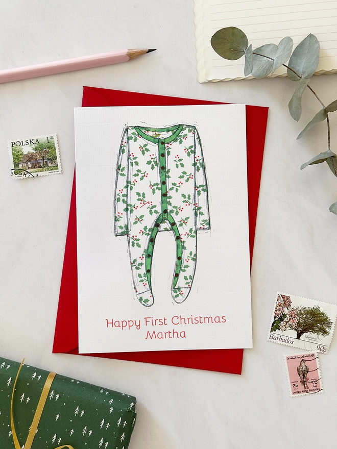 A personalised first Christmas card with an illustrated onesie lays on a red envelope beside various stationery items on a white desk.