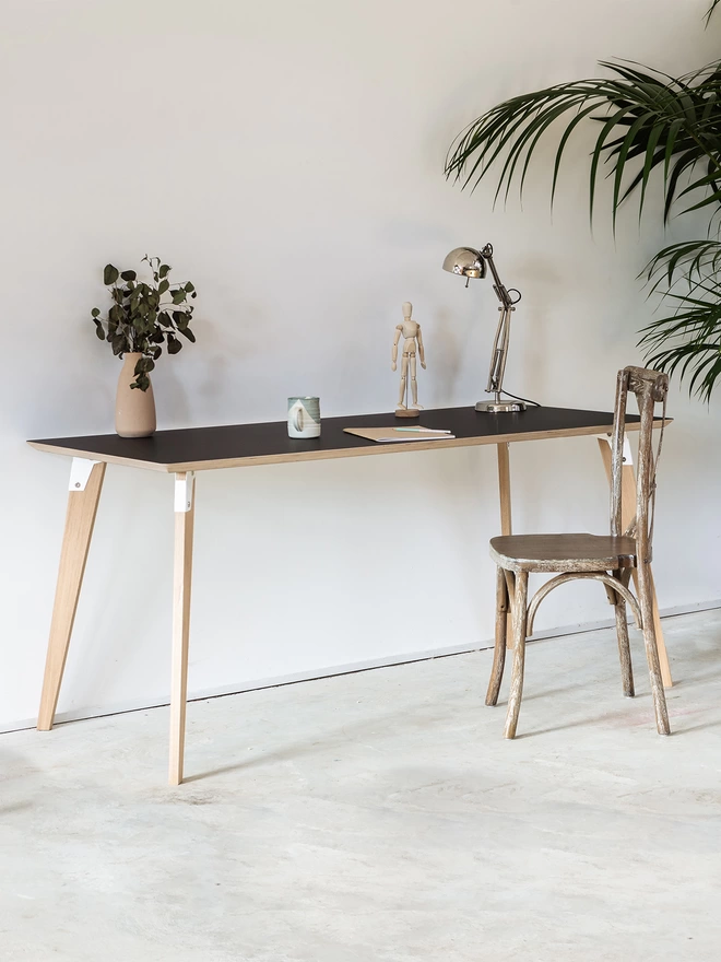 A stylish and minimalist desk with black Fenix top, white coloured steel brackets and solid oak legs, with a chair in front and a plant and light on the desk.