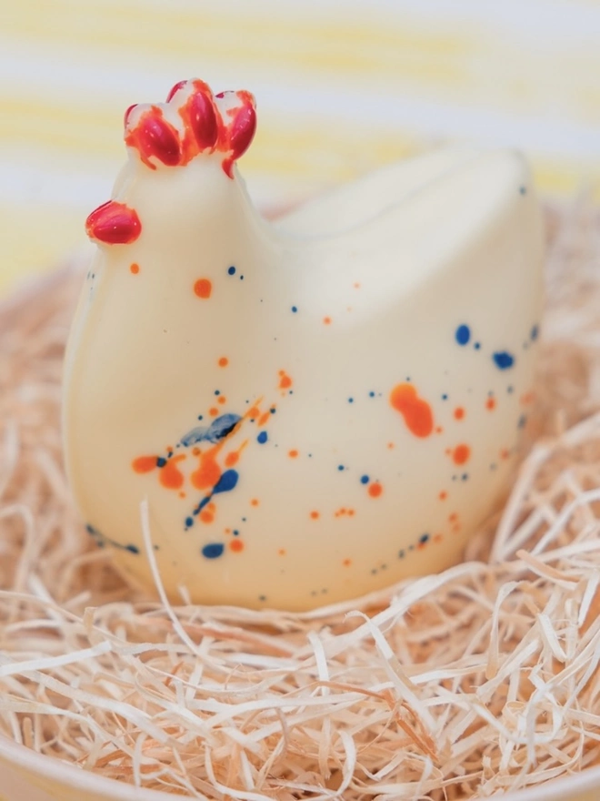 A white chocolate chicken from Nenette Chocolates, sitting in some straw in a yellow bowl on a yellow and white striped tablecloth
