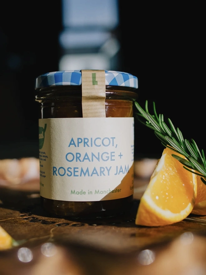 Jar Of Apricot, Orange & Rosemary Jam, jar of jam next to a rosemary leaf and slice of orange on a decorated plate. 