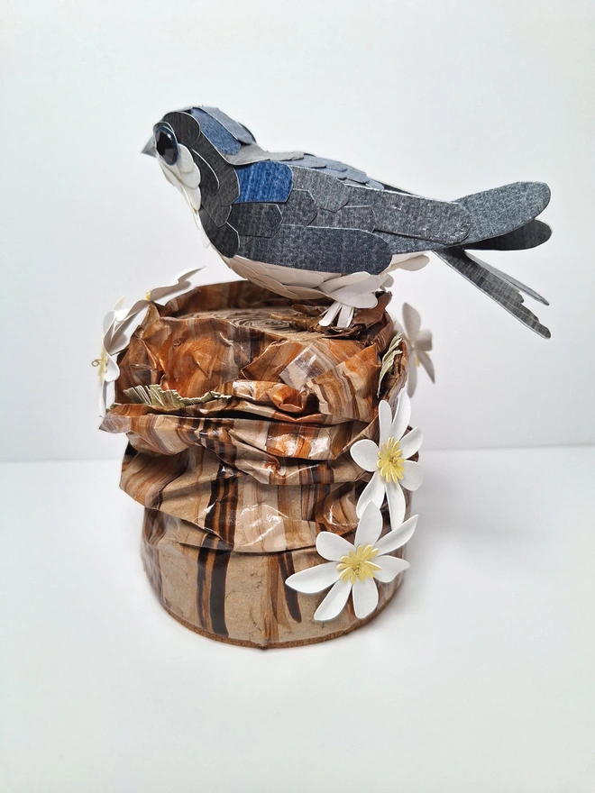 Housemartin bird sculpture, perched on a floral tree stump. This handmade sculpture is made from recycled paper. The bird is black and blue, with a white belly. White and yellow flowers adorn the tree stump. Back view.