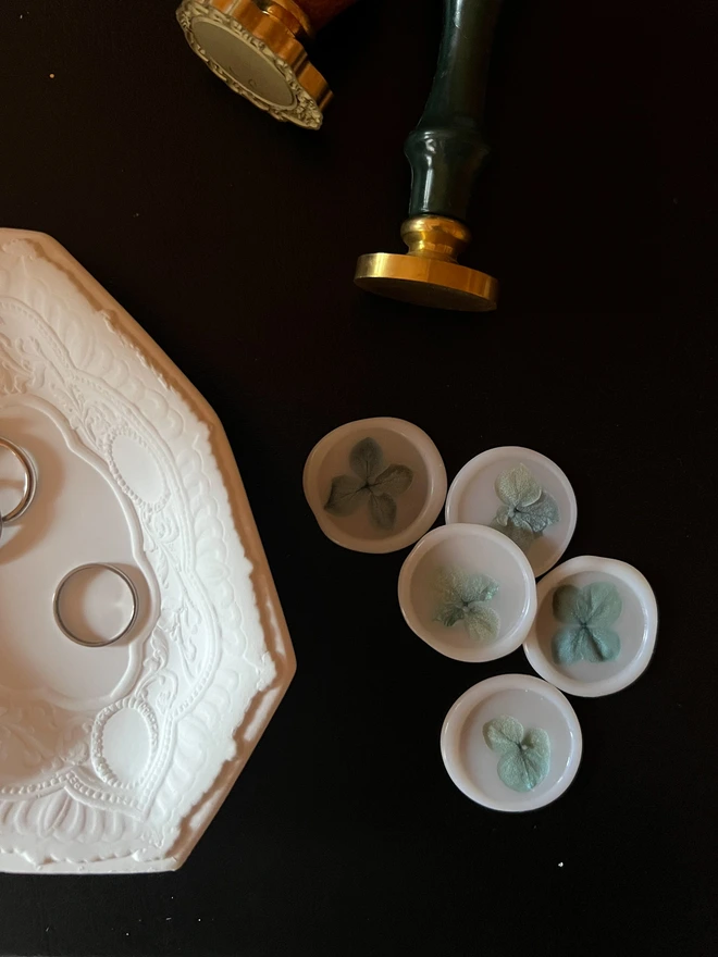 White vellum wax seals with blue hydrangea petals on a black desk, laid with two wax seal stamps and a ceramic trinket tray containing rings 