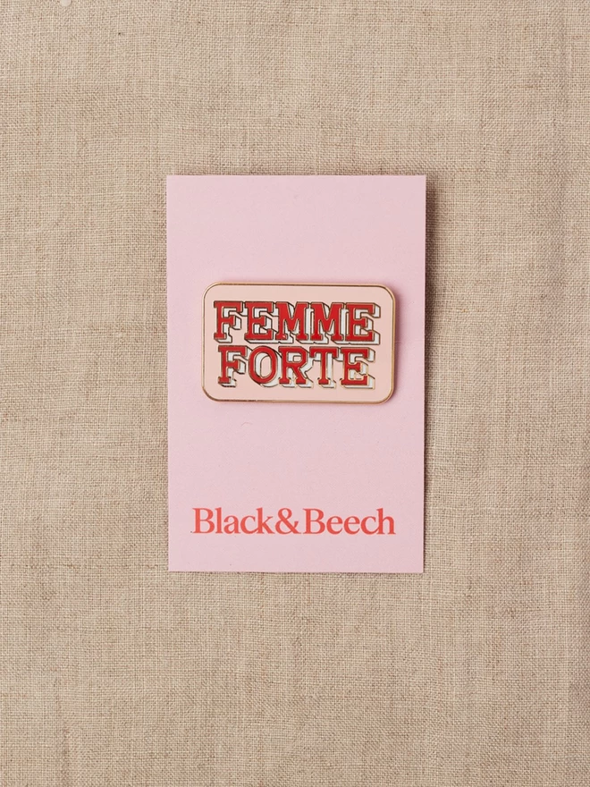 A calamine pink rectangular enamel pin with the words Femme Forte written in red 
