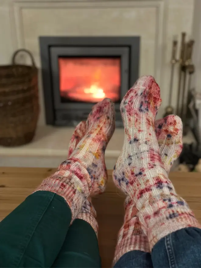 Two pairs of feet wearing studioEVIG's naturally dyed alpaca lounge socks, comfortably propped up on a table in front of a glowing fireplace, evoking warmth and relaxation