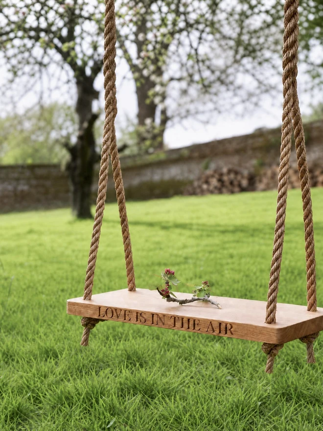 Personalised solid oak swing hanging from rope in a walled orchard, engraved 'Love is in the air'
