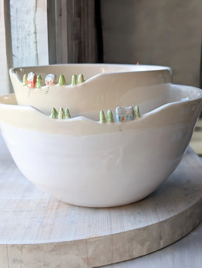 A handmade ceramic white 2 stacked white bowls with tiny houses and tree details on the rim and white glaze  sitting on a wooden surface looking rather gorgeous