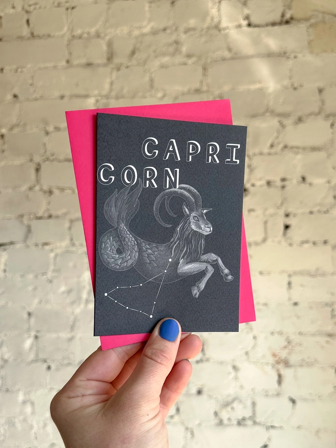a greetings card featuring a greyscale illustration of a zodiac style sea goat with the text “CAPRICORN” with a neon pink envelope